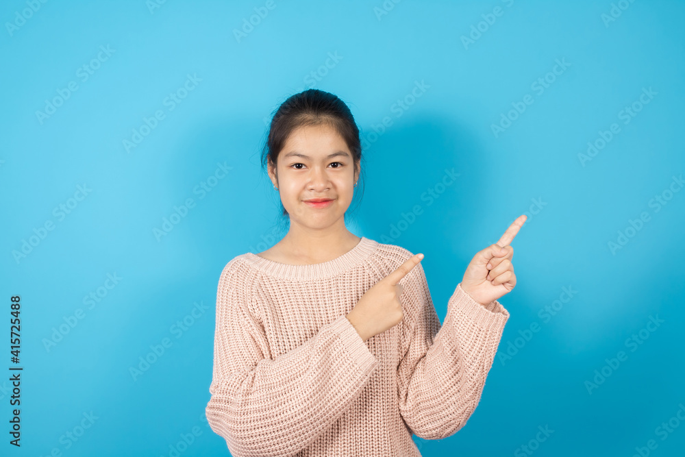 Portrait of smiling Asian teenager joyfully and pointing finger to the side, Wears in light pink knitted sweater isolated over blue background with copy space Portrait of smiling Asian teenager joyfully and pointing finger to the side, Wears in light pink knitted sweater isolated over blue background with copy space