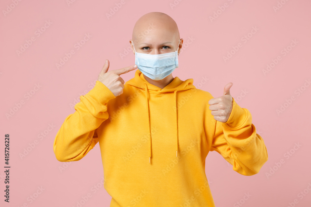 Young bald woman 20s without hair in yellow casual shirt point index finger on sterile face mask to safe from coronavirus covid-19, quarantine show thumb up gesture isolated on pastel pink background Young bald woman 20s without hair in yellow casual shirt point index finger on sterile face mask to safe from coronavirus covid-19, quarantine show thumb up gesture isolated on pastel pink background
