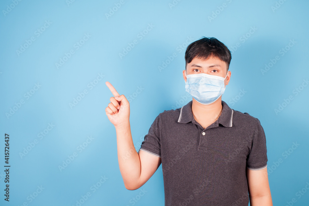 Asian young man wearing face mask protects, air pollution and COVID virus her pointing out side on blue background Asian young man wearing face mask protects, air pollution and COVID virus her pointing out side on blue background