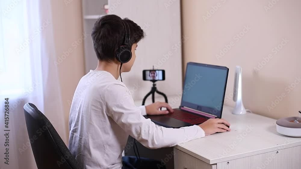 Teenage boy playing game on his pc computer in white room. Gamer capture video on web camera on laptop and phone on tripod Teenage boy playing game on his pc computer in white room. Gamer capture video on web camera on laptop and phone on tripod