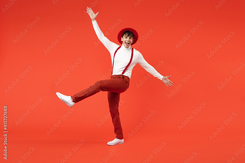 Fototapeta premium Full length of young spanish latinos surprised stylish man 20s in hat white shirt trousers with suspenders leaning over with outstratched hands raised up leg isolated on red background studio portrait