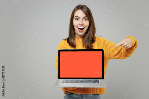 Young freelancer copywriter cute attractive woman 20s in knitted yellow sweater point index finger on laptop pc computer with blank screen workspace area isolated on grey background studio portrait.. photo