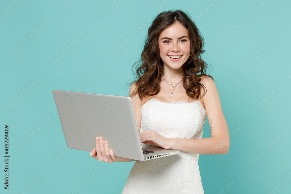 Cheerful funny bride young woman 20s in beautiful white wedding dress working on laptop pc computer isolated on blue turquoise color background studio portrait. Ceremony celebration party concept. Cheerful funny bride young woman 20s in beautiful white wedding dress working on laptop pc computer isolated on blue turquoise color background studio portrait. Ceremony celebration party concept.