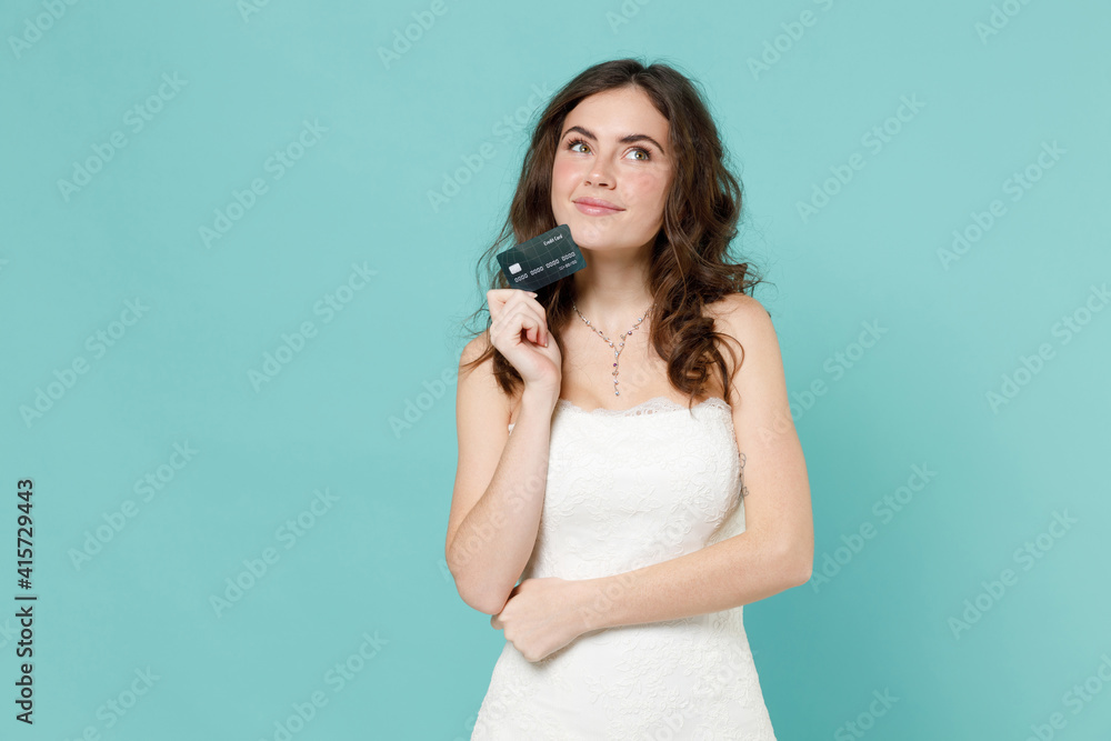 Pensive bride young woman 20s in beautiful white wedding dress hold credit bank card looking aside up isolated on blue turquoise color background studio portrait. Ceremony celebration party concept. Pensive bride young woman 20s in beautiful white wedding dress hold credit bank card looking aside up isolated on blue turquoise color background studio portrait. Ceremony celebration party concept.