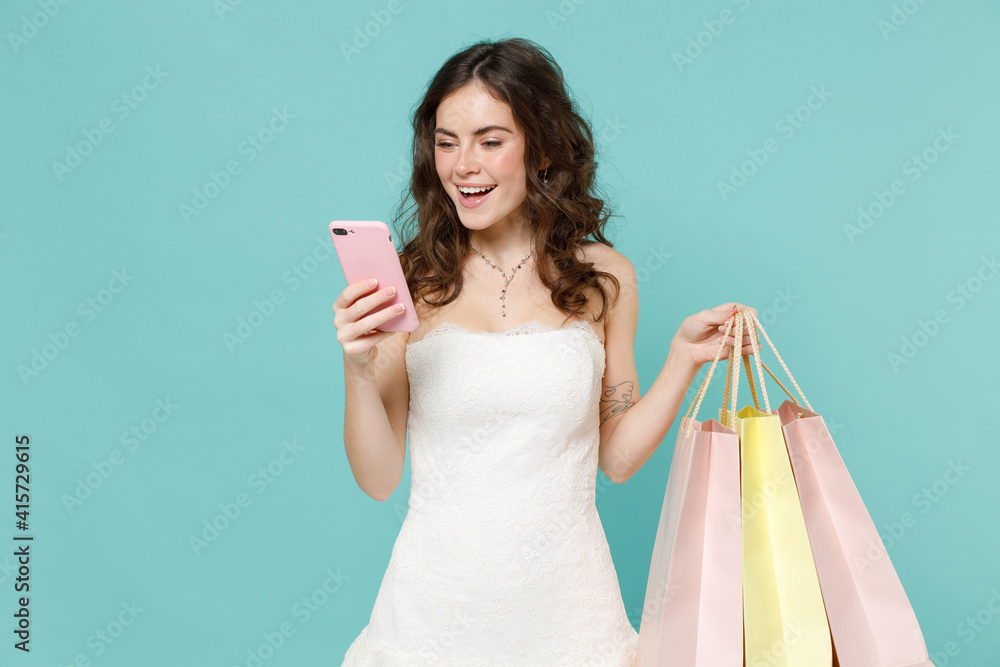 Cheerful bride young woman in white wedding dress hold package bags with purchases after shopping using mobile phone isolated on blue turquoise background studio. Ceremony celebration party concept. Cheerful bride young woman in white wedding dress hold package bags with purchases after shopping using mobile phone isolated on blue turquoise background studio. Ceremony celebration party concept.