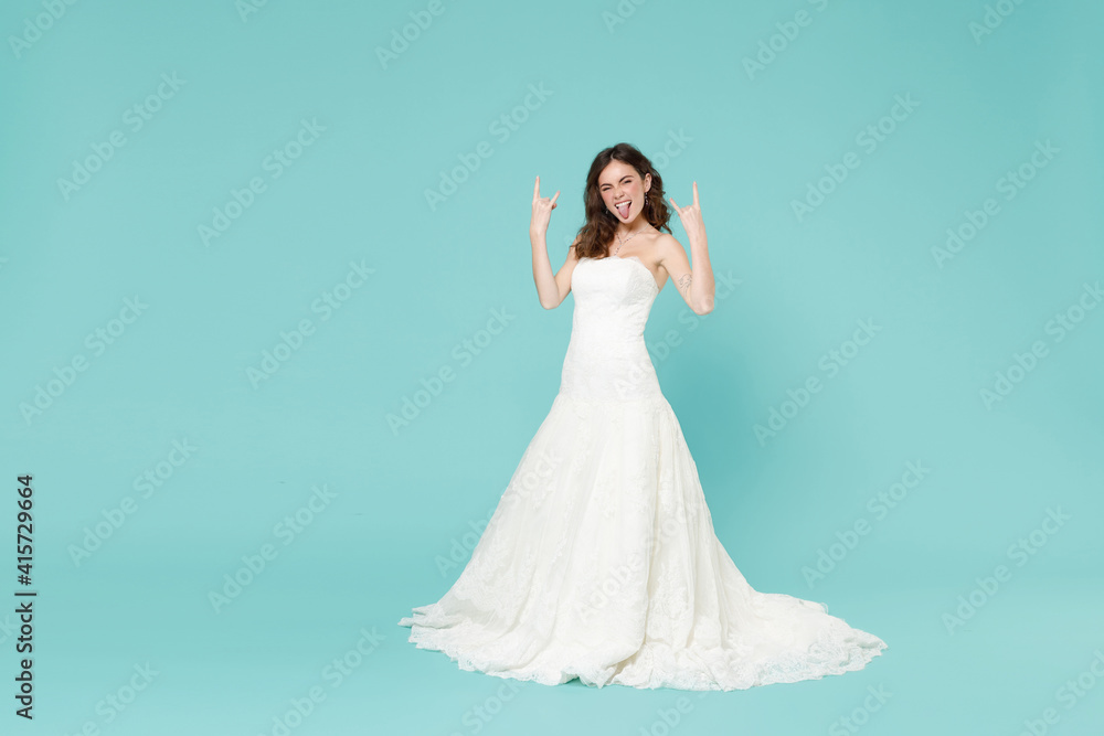 Full length cheerful bride young woman 20s in white wedding dress depicting heavy metal rock sign horns up gesture isolated on blue turquoise background studio. Ceremony celebration party concept. Full length cheerful bride young woman 20s in white wedding dress depicting heavy metal rock sign horns up gesture isolated on blue turquoise background studio. Ceremony celebration party concept.