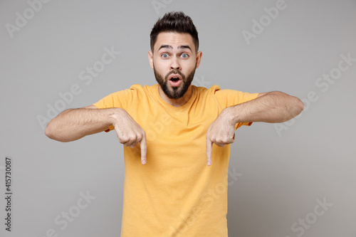Young caucasian bearded surprised shocked man in yellow basic t-shirt point index finger down on workspace area copy space mock up isolated on grey background studio portrait People lifestyle concept.