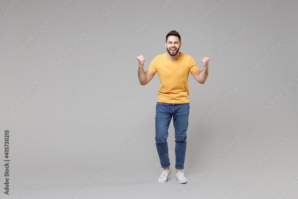 Full length young caucasian serious bearded attractive handsome man 20s wear casual yellow basic t-shirt do winner gesture clench fist isolated on grey color background studio portrait People concept. Full length young caucasian serious bearded attractive handsome man 20s wear casual yellow basic t-shirt do winner gesture clench fist isolated on grey color background studio portrait People concept.