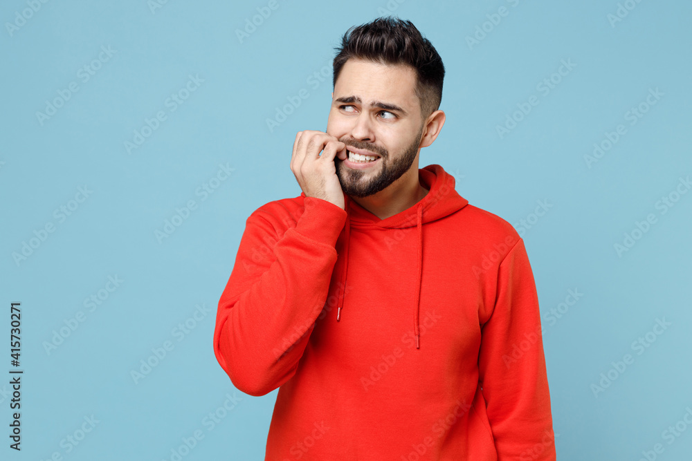 Fototapeta premium Young caucasian troubled pensive stressed worried bearded man 20s in casual red orange hoodie looking aside biting nails finger isolated on blue background studio portrait People lifestyle concept.