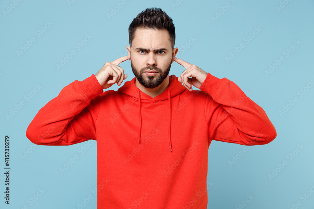 Young caucasian angry nervous irritated stressed bearded man 20s in casual red orange hoodie covering ears with hand fingers isolated on blue color background studio portrait People lifestyle concept. Young caucasian angry nervous irritated stressed bearded man 20s in casual red orange hoodie covering ears with hand fingers isolated on blue color background studio portrait People lifestyle concept.