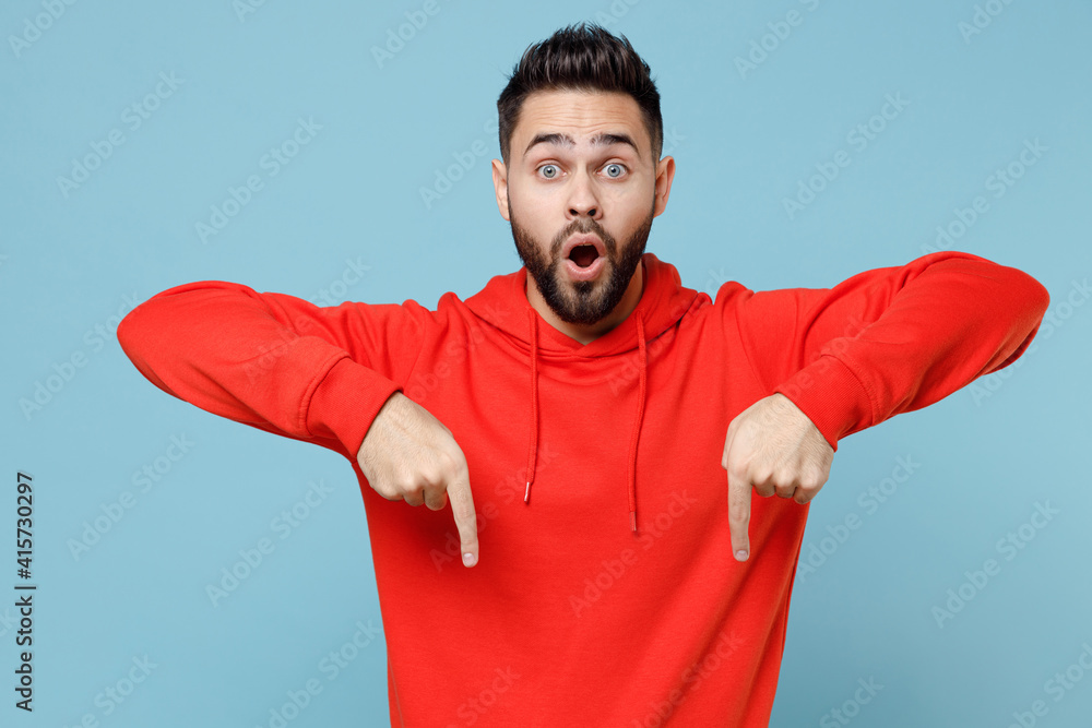 Fototapeta premium Young caucasian smiling bearded attractive man in casual red orange hoodie point index finger overhead on copy space area mock up isolated on blue background studio portrait People lifestyle concept.