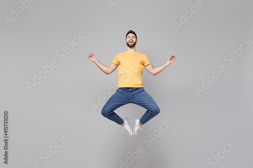 Full length of young bearded man 20s wearing yellow basic t-shirt jump high hold hands in yoga gesture, relaxing meditating, trying to calm down levitating isolated on grey background studio portrait