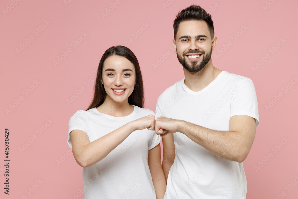Young cheerful couple two friends man woman 20s in white basic t-shirts giving fist bump holding hands folded hit bang gesture isolated on pastel pink background Friendship greet meet hello hi salute. Young cheerful couple two friends man woman 20s in white basic t-shirts giving fist bump holding hands folded hit bang gesture isolated on pastel pink background Friendship greet meet hello hi salute.