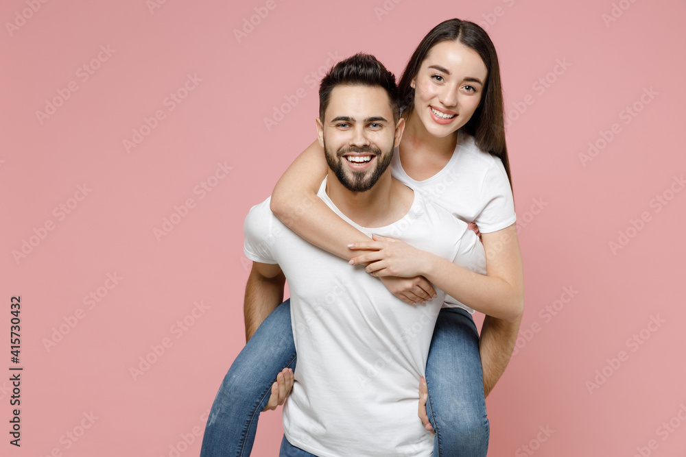Young cheerful lovely couple two friends man woman in white basic blank t-shirts giving piggyback ride to joyful sits on back fooling around isolated on pastel pink color background studio portrait Young cheerful lovely couple two friends man woman in white basic blank t-shirts giving piggyback ride to joyful sits on back fooling around isolated on pastel pink color background studio portrait