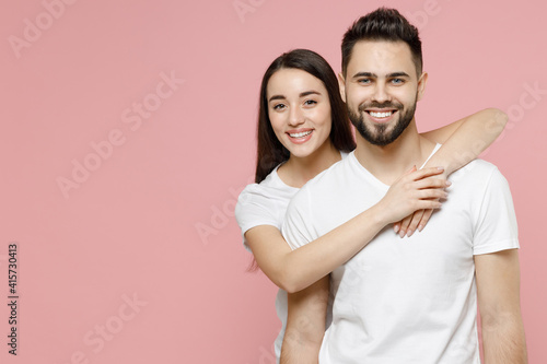 Young cheerful happy couple two friends bearded man brunette woman in white basic blank print design t-shirt standing behind hug hands embrace isolated on pastel pink color background studio portrait