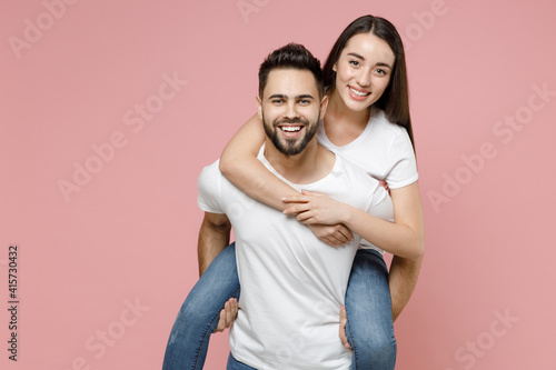 Young cheerful lovely couple two friends man woman in white basic blank t-shirts giving piggyback ride to joyful sits on back fooling around isolated on pastel pink color background studio portrait