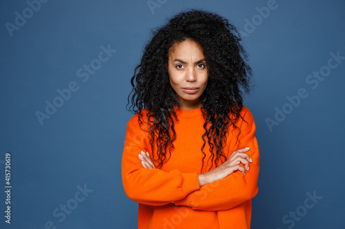 Displeased dissatisfied worried young african american woman wearing casual basic bright orange sweatshirt standing hold hands crossed looking camera isolated on blue color background studio portrait. © ViDi Studio