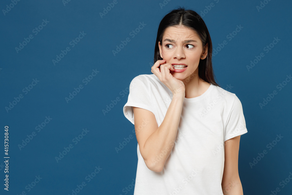 Young troubled thoughtful pensive worrying scared mistaken latin woman 20s wearing white casual basic t-shirt biting nails fingers look aside isolated on dark blue color background studio portrait. Young troubled thoughtful pensive worrying scared mistaken latin woman 20s wearing white casual basic t-shirt biting nails fingers look aside isolated on dark blue color background studio portrait.