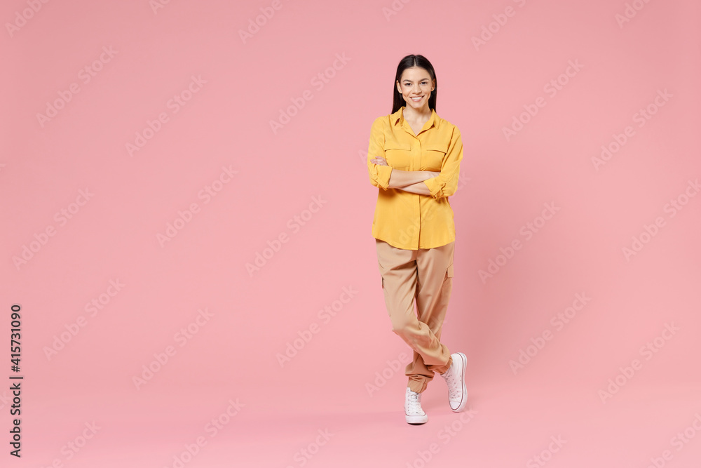 Full length of young smiling pretty happy brunette positive attractive beautiful latin woman 20s in yellow casual shirt hold hands crossed folded isolated on pastel pink background studio portrait. - obrazy, fototapety, plakaty 