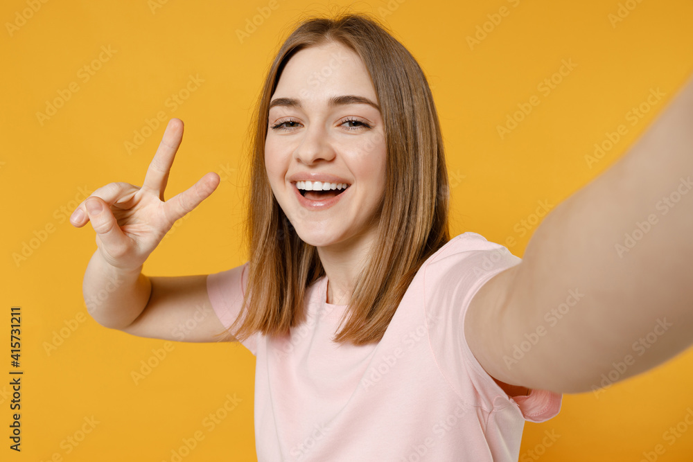 Close up young happy woman 20s with nude make up wearing casual basic pastel pink t-shirt doing selfie shot on mobile phone show victory v-sign gesture isolated on yellow background studio portrait Close up young happy woman 20s with nude make up wearing casual basic pastel pink t-shirt doing selfie shot on mobile phone show victory v-sign gesture isolated on yellow background studio portrait