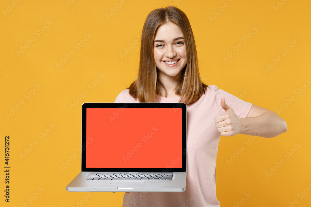 Naklejka premium Young freelancer woman 20s in casual basic pastel pink t-shirt holding laptop pc computer with blank screen workspace area show thumb up like gesture isolated on yellow background studio portrait