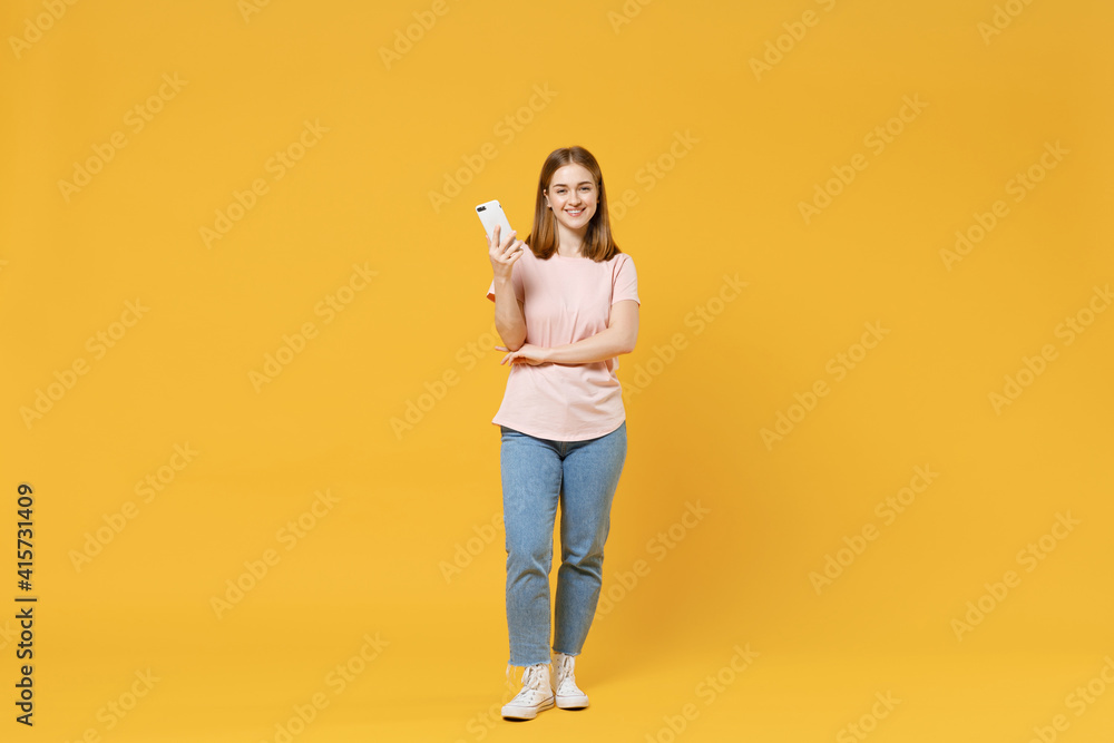 Full length of young caucasian student woman 20s wearing casual basic pastel pink t-shirt, jeans holding mobile cell phone chatting surfing internet isolated on yellow background studio portrait. Full length of young caucasian student woman 20s wearing casual basic pastel pink t-shirt, jeans holding mobile cell phone chatting surfing internet isolated on yellow background studio portrait.