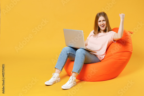 Full length of young woman 20s in basic pastel pink t-shirt, jeans sitting in orange bean bag chair holding laptop pc computer do winner gesture clench fist celebrating isolated on yellow background. © ViDi Studio