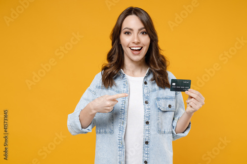 Young surprised happy excited rich successful positive woman 20s in stylish denim shirt white t-shirt point index finger on credit bank card look camera isolated on yellow background studio portrait