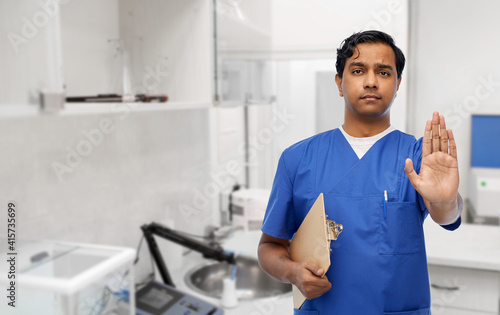 healthcare, profession and medicine concept - indian male doctor in blue uniform with clipboard showing stop gesture over laboratory or hospital background