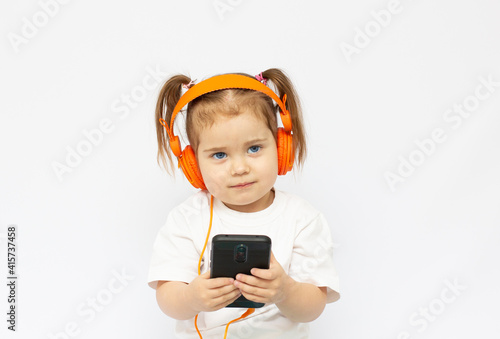 The little girl in headphones. The girl is listening music. The little girl is playing phone on the background