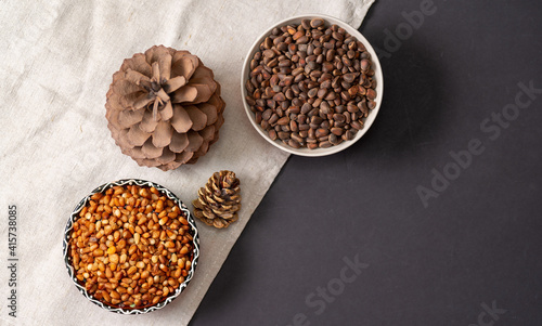 Two curly bowls with peeled pine nuts and nuts, two pine cones on a linen cloth and a black table. 