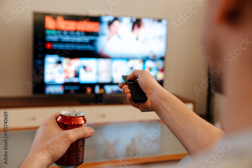 Caucasian guy drinking soda and using remote control while watching TV