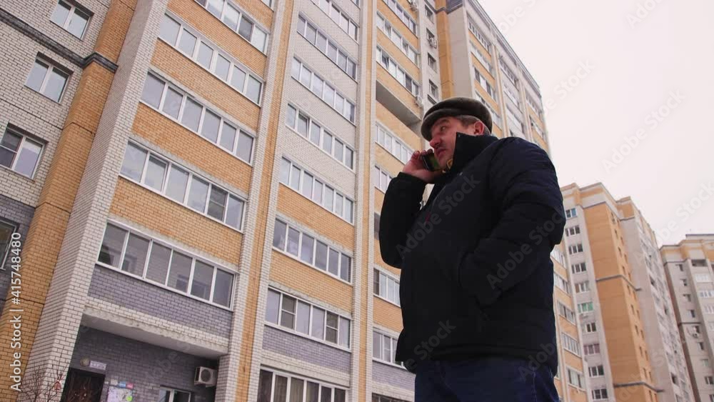 A man stands in the yard, waiting for a taxi and talking on the phone. Male with a mustache in a warm jacket outdoors in cool weather. In the background, multi-storey high-rise buildings out of focus A man stands in the yard, waiting for a taxi and talking on the phone. Male with a mustache in a warm jacket outdoors in cool weather. In the background, multi-storey high-rise buildings out of focus
