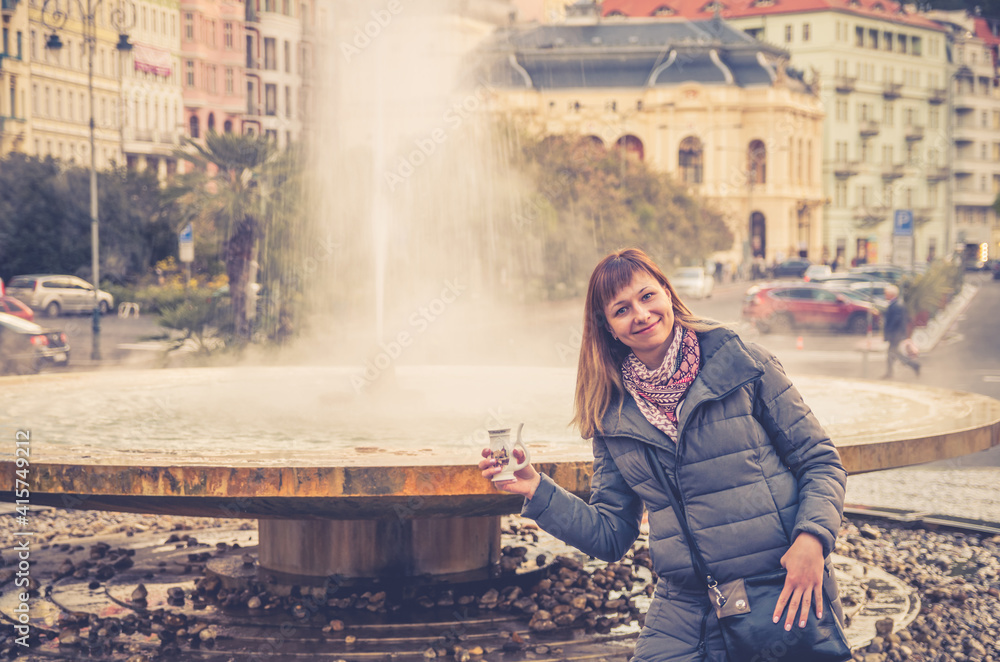 Fototapeta premium Young woman tourist with grey jacket posing and hold therapeutic mineral water in cup near fountain hot spring geyser Vridlo in Karlovy Vary historical city centre, girl on vacation in Czech Republic