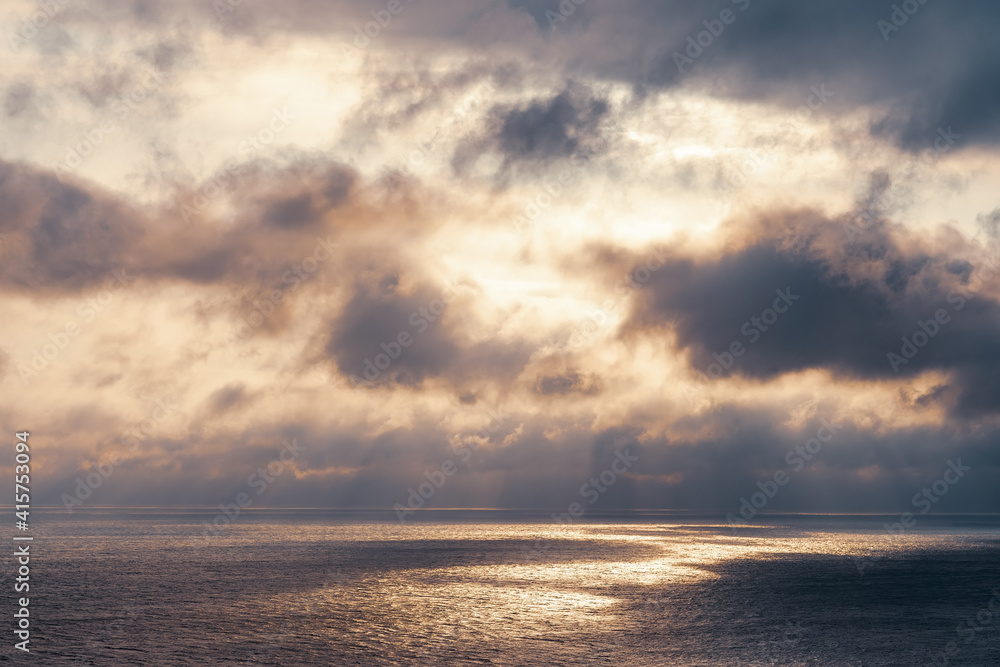The sun's rays make their way through dramatic dark clouds over the sea, patches of light on the water