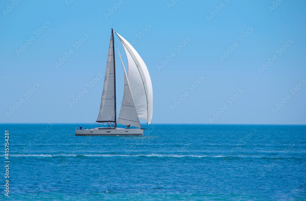 Sailboat with white sails on the open sea against the backdrop of a clear blue sky.