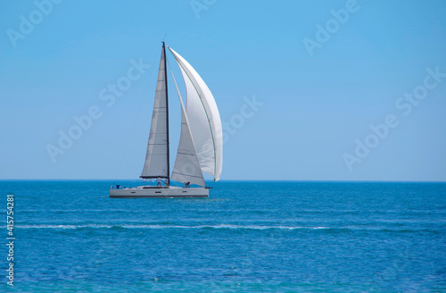 Sailboat with white sails on the open sea against the backdrop of a clear blue sky.