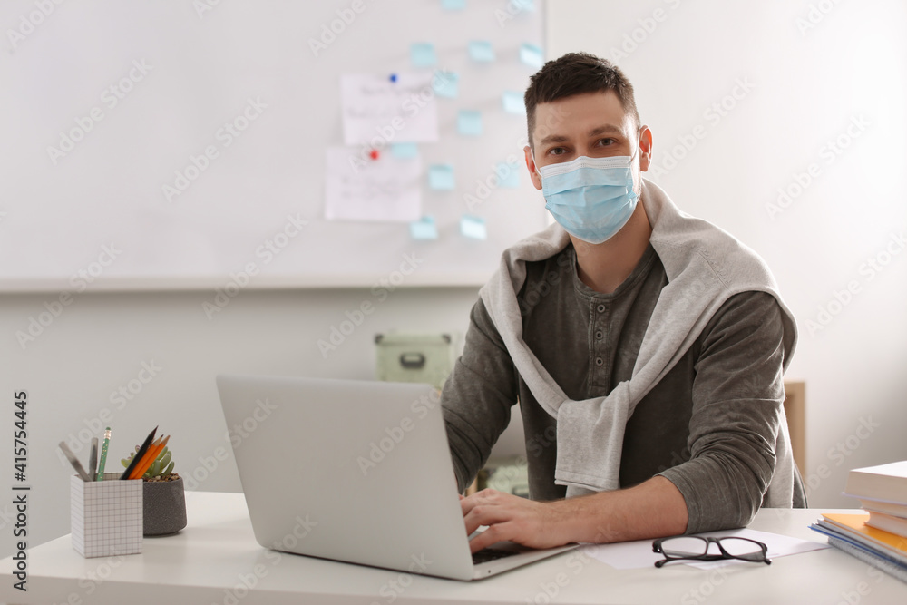 Teacher with protective mask and laptop sitting at desk in classroom. Reopening after Covid-19 quarantine Teacher with protective mask and laptop sitting at desk in classroom. Reopening after Covid-19 quarantine