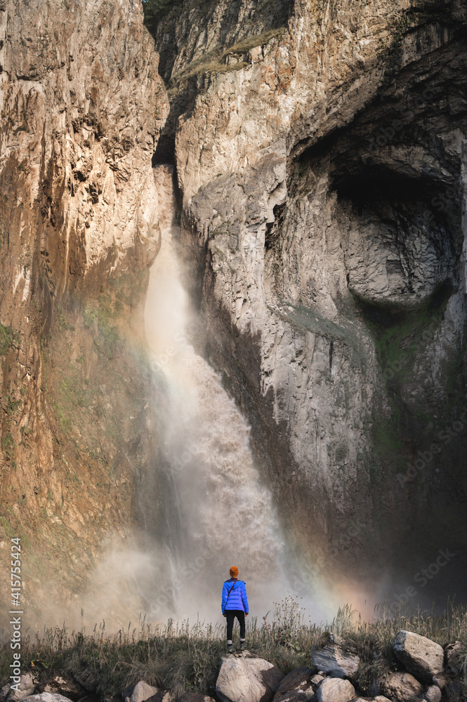 Obraz premium A woman in a warm jacket and hat stands with her back against the background of a huge epic waterfall flowing from large mountain rocks
