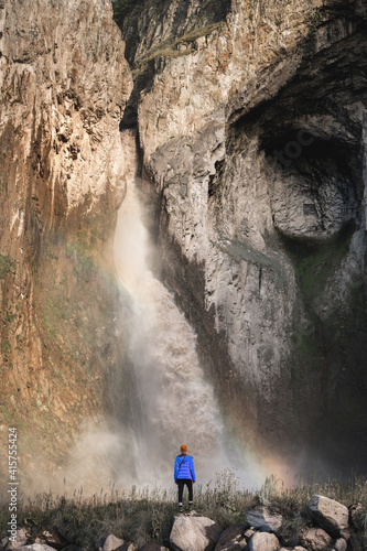 A woman in a warm jacket and hat stands with her back against the background of a huge epic waterfall flowing from large mountain rocks