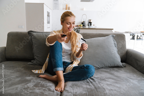 Happy woman holding credit card and watching tv with remote control