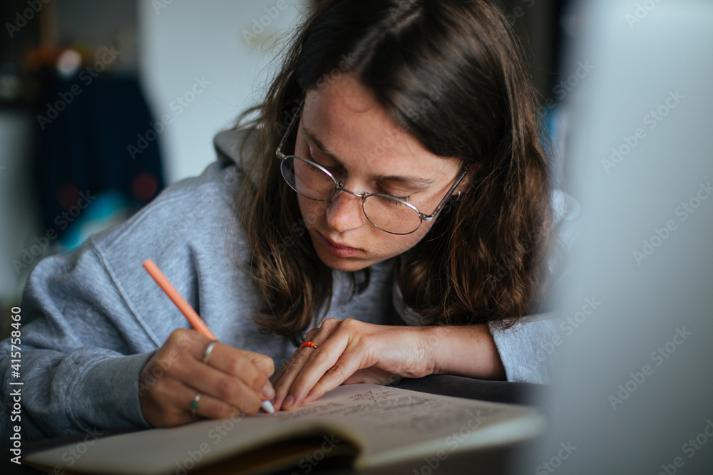 Cinematic shot of young female make notes in notebook with pen, look at laptop. Millennial woman use free time to learn new skill from online courses. E-learning and new technology. Home schooling Cinematic shot of young female make notes in notebook with pen, look at laptop. Millennial woman use free time to learn new skill from online courses. E-learning and new technology. Home schooling