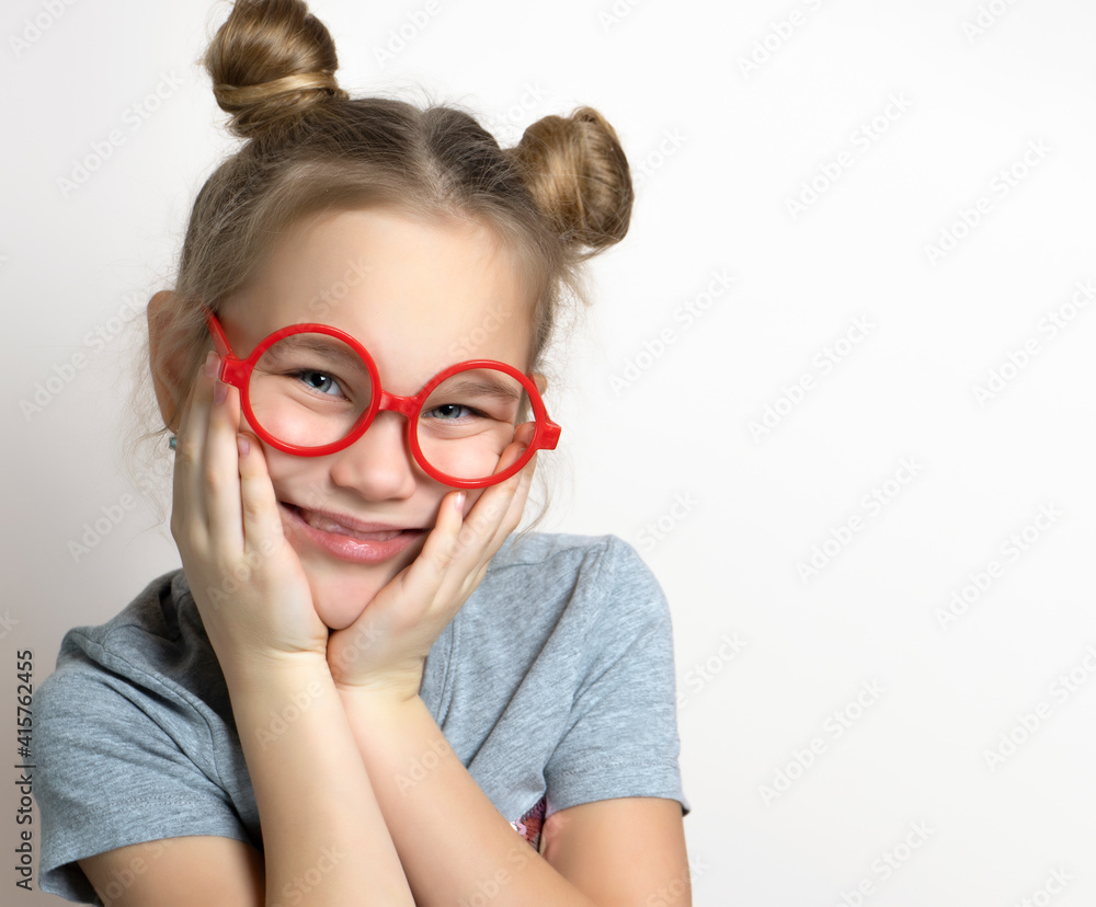 Cute funny little girl with smiling facial emotion wearing red toy eyeglasses touching cheeks studio headshot portrait isolated on grey. Beautiful lovely caucasian female child with cute bun hairstyle Cute funny little girl with smiling facial emotion wearing red toy eyeglasses touching cheeks studio headshot portrait isolated on grey. Beautiful lovely caucasian female child with cute bun hairstyle