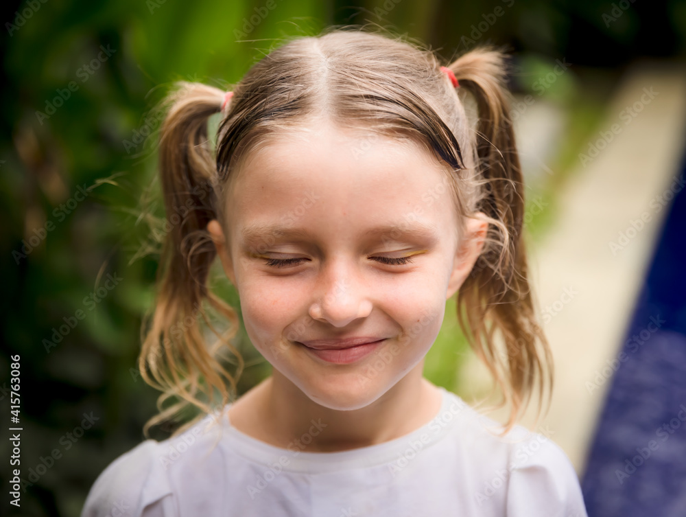 Close up portrait of pretty little girl. Face with closed eyes. Caucasian girl with two ponytails. Green tropical leaves and swimming pool background.