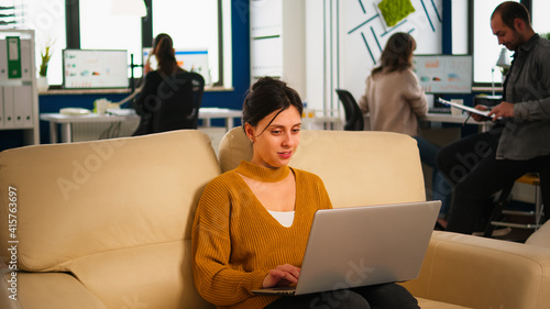 Happy business woman reading good news on laptop smiling, sitting on couch in busy start up company office, while diverse team analyzes statistics datas. Multiethnic team talking about project. © DC Studio