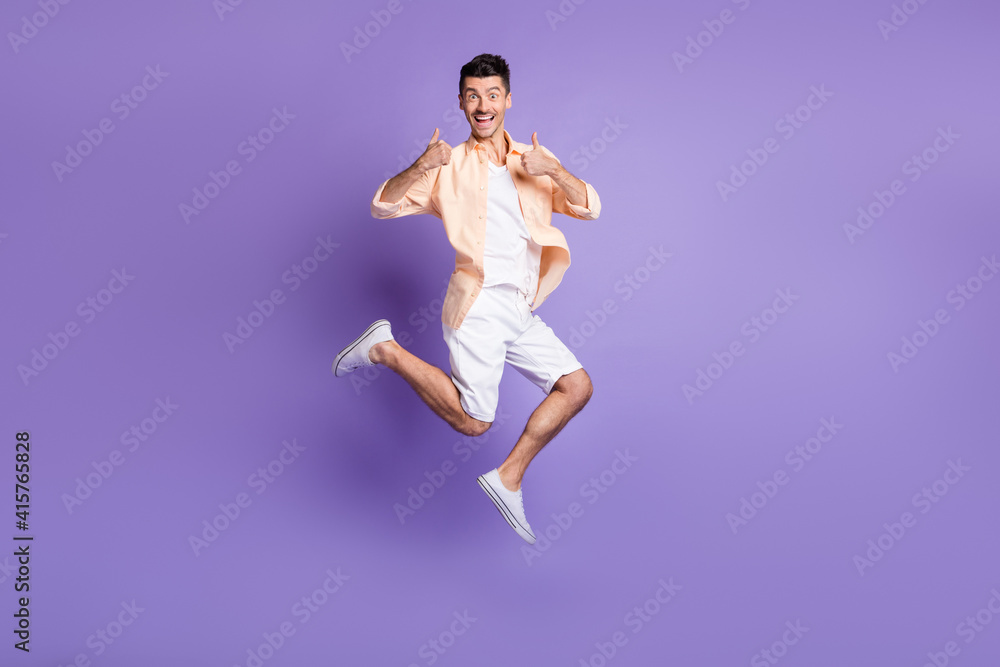 Full length photo of man jump raise two thumbs up wear beige shirt shorts sneakers isolated purple color background