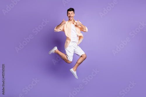 Full length photo of man jump raise two thumbs up wear beige shirt shorts sneakers isolated purple color background
