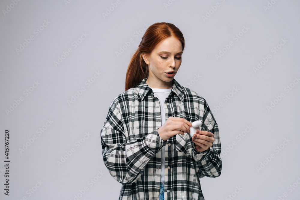 Attractive young woman taking wireless earphones out of case on isolated white background. Pretty redhead lady model wearing casual fashion clothes emotionally showing facial expressions, copy space. Attractive young woman taking wireless earphones out of case on isolated white background. Pretty redhead lady model wearing casual fashion clothes emotionally showing facial expressions, copy space.