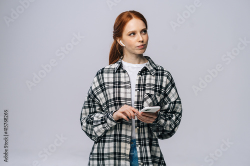 Thinking red-haired young woman wearing wireless earphones typing online message on isolated white background, looking away. Pretty redhead lady model emotionally showing facial expressions.