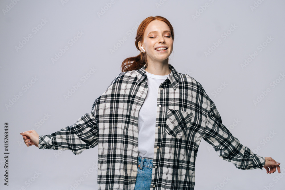 Joyful young woman wearing wireless earphones dancing while listening popular song on isolated white background, closed eyes. Pretty redhead lady model emotionally showing facial expressions. Joyful young woman wearing wireless earphones dancing while listening popular song on isolated white background, closed eyes. Pretty redhead lady model emotionally showing facial expressions.